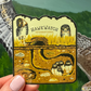 A hand holds a sticker featuring a Burrowing Owl illustration and the text 'HAWKWATCH INTERNATIONAL' 